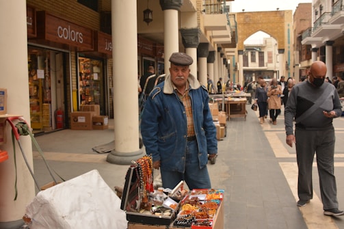 A street vendor wearing a denim jacket and cap stands near a stall displaying an assortment of colorful necklaces and small items. The setting is a marketplace lined with tall, white columns and several people walking along the walkway. A shop named 'Colors' is visible in the background alongside various store signs and open storefronts.