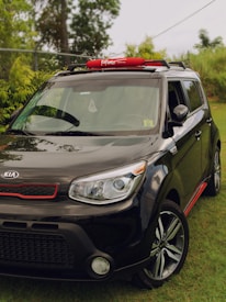 A black Kia Soul car is parked on a grassy area. It has a red rooftop cargo carrier labeled 'Komoto', and red accents around the front grille and side skirts. The car is positioned at an angle, with trees and a fence visible in the background.