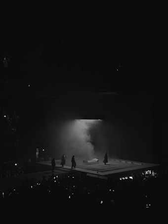 A dark stage featuring a rectangular platform with people dressed in dark clothing walking towards a white couch or bench. The scene is enveloped in mist or smoke, creating a dramatic atmosphere. An audience can be seen in the dimly lit foreground.