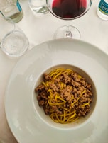 A bowl of pasta with minced meat sauce is set on a dining table. Nearby, there is a glass of red wine and a few sparkling water bottles, suggesting a fine dining experience.