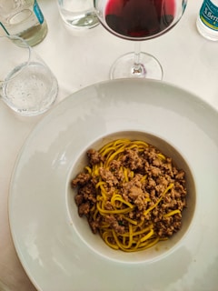 A bowl of pasta with minced meat sauce is set on a dining table. Nearby, there is a glass of red wine and a few sparkling water bottles, suggesting a fine dining experience.