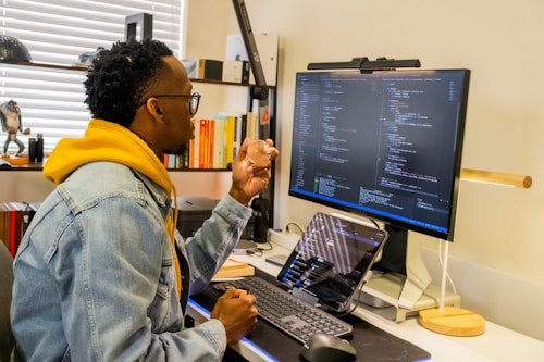A person with short, curly hair and glasses is sitting at a desk, writing code on a large monitor. Wearing a denim jacket over a yellow hoodie, they are focused on the screen displaying multiple lines of code. The desk is organized with a keyboard, mouse, tablet, and various books in the background.