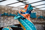 A vibrant sculpture with swirling patterns and vivid colors stands prominently in front of a modern architectural background. The sculpture is adorned with orange, blue, and teal colors, with polka dot accents. Behind it, there is signage with stylized text, suggesting a cultural theme or venue.