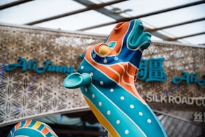 A vibrant sculpture with swirling patterns and vivid colors stands prominently in front of a modern architectural background. The sculpture is adorned with orange, blue, and teal colors, with polka dot accents. Behind it, there is signage with stylized text, suggesting a cultural theme or venue.