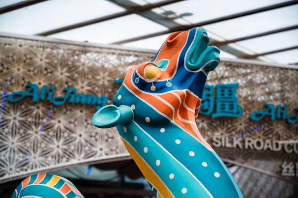 A vibrant sculpture with swirling patterns and vivid colors stands prominently in front of a modern architectural background. The sculpture is adorned with orange, blue, and teal colors, with polka dot accents. Behind it, there is signage with stylized text, suggesting a cultural theme or venue.