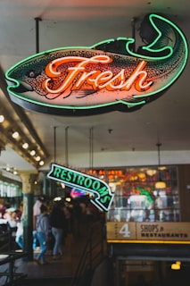 Neon signs hang in an indoor market area, one shaped like a fish with the word 'Fresh' and another pointing to 'Restrooms.' The background shows a bustling environment with people walking and illuminated signs for shops and restaurants.