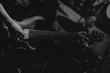 A person is playing an electric guitar, with a focus on the hands and the guitar's body. The image is in black and white, providing a dramatic and intense atmosphere. In the background, there is a blurred figure, possibly a drummer.