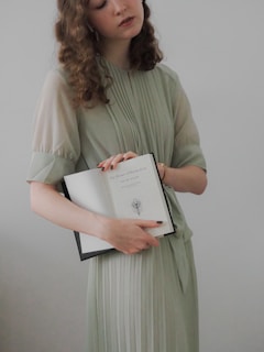 A person with curly hair is wearing a light green, pleated dress and holding an open book titled 'The Picture of Dorian Gray' by Oscar Wilde. The cover page includes a simple illustration of a peacock feather.