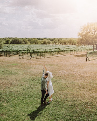 A couple is dancing in a sunlit vineyard. The bride is wearing a white dress and veil, while the groom is in a casual shirt. They are surrounded by vast green fields and rows of grapevines. A large tree stands in the distance under a partly cloudy sky.