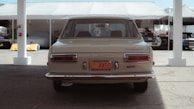 A vintage car is parked under a tent with other vehicles visible in the background. The rear view highlights its classic design and historical vehicle license plate. The scene has a soft lighting with shadows cast underneath the tent structure.