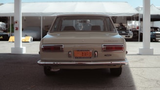 A vintage car is parked under a tent with other vehicles visible in the background. The rear view highlights its classic design and historical vehicle license plate. The scene has a soft lighting with shadows cast underneath the tent structure.