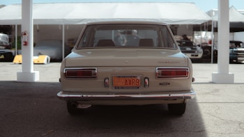 A vintage car is parked under a tent with other vehicles visible in the background. The rear view highlights its classic design and historical vehicle license plate. The scene has a soft lighting with shadows cast underneath the tent structure.
