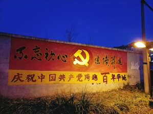 A mural on a wall with Chinese text and the prominent symbol of the hammer and sickle against a red background. The mural is illuminated by a nearby streetlight, and the sky is deep blue. The ground in front of the mural is grassy.