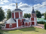 A red church with white trim and a black metal roof, featuring a central dome topped with a golden spire. The building is set within a manicured garden with blooming flowers and surrounded by green trees. The sky is bright with fluffy white clouds.