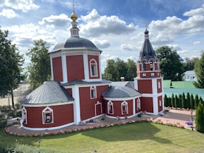 A red church with white trim and a black metal roof, featuring a central dome topped with a golden spire. The building is set within a manicured garden with blooming flowers and surrounded by green trees. The sky is bright with fluffy white clouds.