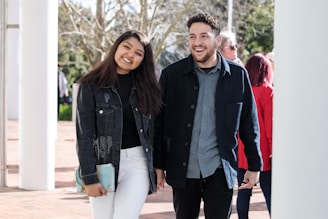 A young woman and man are walking together outdoors, smiling warmly. The woman is wearing a dark denim jacket over a black top with white pants, holding a clutch. The man is dressed in a dark jacket over a button-up shirt with black pants. There are trees and several other people in the background, suggesting a casual, sunny day.