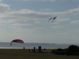 A serene coastal scene with a large red and blue kite in the foreground and another kite soaring higher in the sky. Several people are gathered near the shoreline, enjoying the activity on a grassy expanse. The sea is calm and stretches toward the horizon under a sky dotted with fluffy clouds.