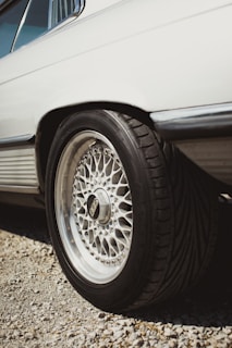 The image focuses on the rear side of a vehicle, highlighting its intricate alloy wheel design. The tire is thick and appears to be designed for performance. The body of the vehicle is clean and white, and the scene appears to be outdoors with gravel under the car.