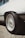 The image focuses on the rear side of a vehicle, highlighting its intricate alloy wheel design. The tire is thick and appears to be designed for performance. The body of the vehicle is clean and white, and the scene appears to be outdoors with gravel under the car.