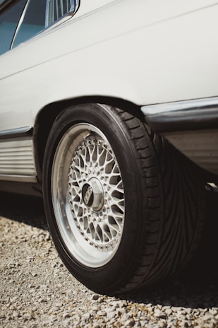 The image focuses on the rear side of a vehicle, highlighting its intricate alloy wheel design. The tire is thick and appears to be designed for performance. The body of the vehicle is clean and white, and the scene appears to be outdoors with gravel under the car.