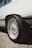 The image focuses on the rear side of a vehicle, highlighting its intricate alloy wheel design. The tire is thick and appears to be designed for performance. The body of the vehicle is clean and white, and the scene appears to be outdoors with gravel under the car.