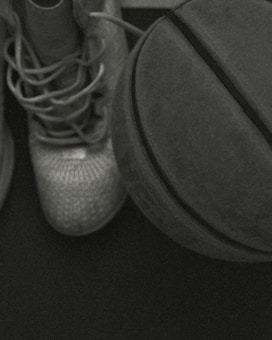 A pair of basketball shoes is situated next to a basketball on a dark surface. The image is in a greenish monochrome tone, creating a vintage or night-vision effect. The laces of the shoes are untied and casually placed, suggesting a casual or rested state.