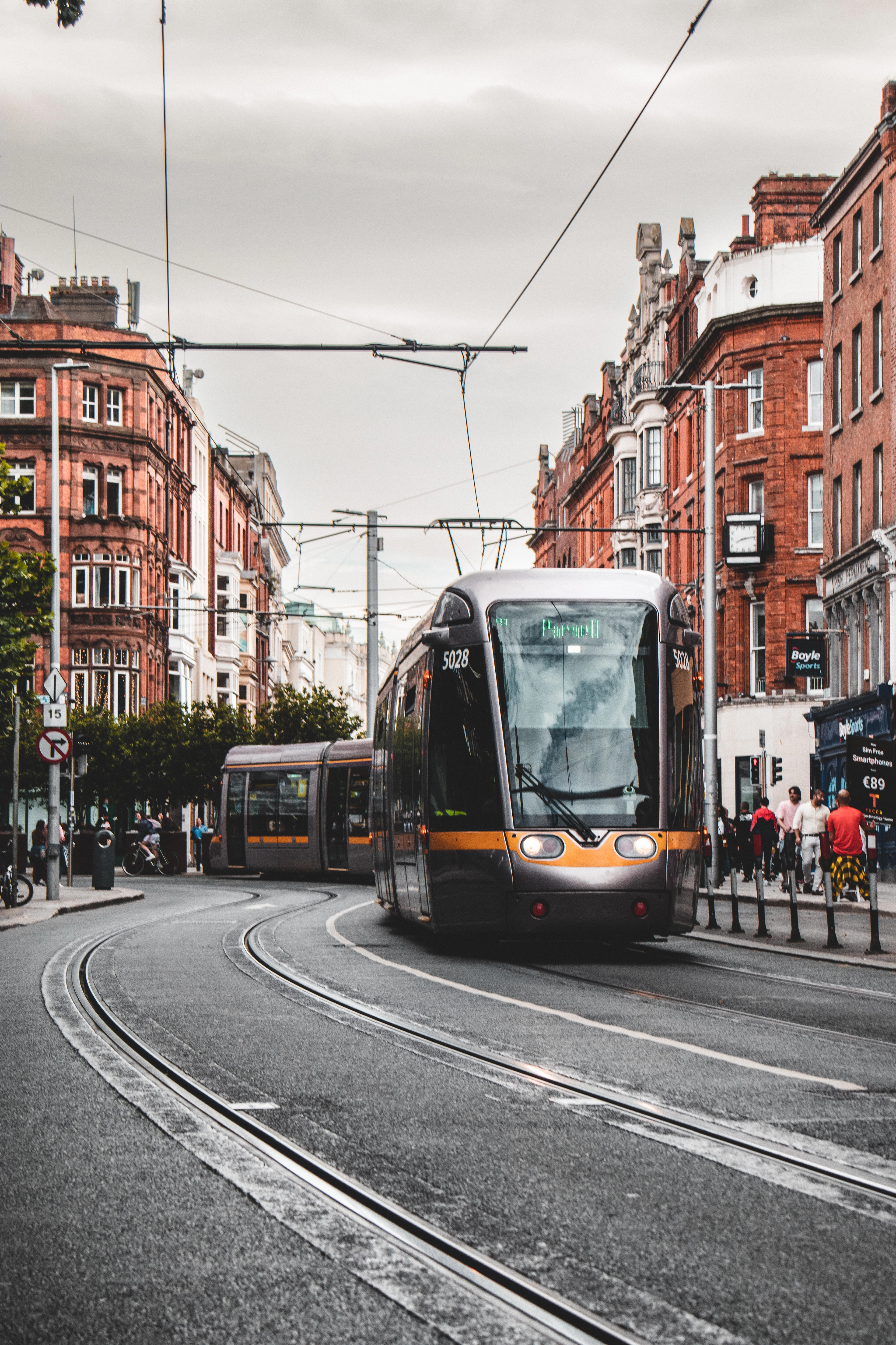 A Luas tram signed for Parnell curving through a Dublin street between red-brick Victorian buildings, a second tram waiting behind it and overhead wires crossing the pale sky