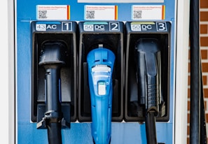 Three different types of electric vehicle charging connectors are mounted on a blue charging station. The connectors are labeled as AC, CHAdeMO, and CCS, each with an associated power output in kW. The station features instructions above the connectors with QR codes.