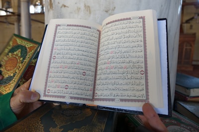 An open book is being held by a person. The book contains Arabic text with decorative borders in red and black ink. Other similar books with ornate covers are positioned nearby on a surface. The setting appears to be a mosque or library, indicated by elements like columns and architectural details in the background.