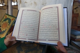 An open book is being held by a person. The book contains Arabic text with decorative borders in red and black ink. Other similar books with ornate covers are positioned nearby on a surface. The setting appears to be a mosque or library, indicated by elements like columns and architectural details in the background.