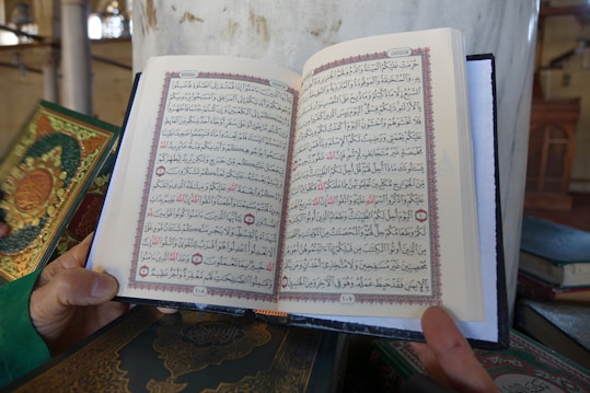 An open book is being held by a person. The book contains Arabic text with decorative borders in red and black ink. Other similar books with ornate covers are positioned nearby on a surface. The setting appears to be a mosque or library, indicated by elements like columns and architectural details in the background.