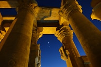 Massive ancient columns with intricate carvings tower upwards against a deep blue night sky. A crescent moon and a bright planet are visible between the columns, adding a celestial element to the scene.