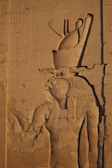 Ancient carved relief depicting a figure with intricate headdress and traditional attire, showcased on a stone wall with hieroglyphics inscribed alongside.