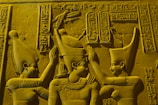 Three figures in traditional ancient Egyptian attire are depicted in a stone carving. The central figure appears to be receiving a ritualistic gesture from the others. Hieroglyphics are present in the background, adding detail and context. The figures wear distinct headpieces and jewelry, indicating their significance.