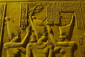 Three figures in traditional ancient Egyptian attire are depicted in a stone carving. The central figure appears to be receiving a ritualistic gesture from the others. Hieroglyphics are present in the background, adding detail and context. The figures wear distinct headpieces and jewelry, indicating their significance.