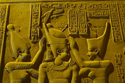 Three figures in traditional ancient Egyptian attire are depicted in a stone carving. The central figure appears to be receiving a ritualistic gesture from the others. Hieroglyphics are present in the background, adding detail and context. The figures wear distinct headpieces and jewelry, indicating their significance.