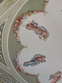 A beautifully painted ceiling features classical figures in flowing garments surrounded by ornate floral arrangements. The figures are depicted in dynamic poses, seemingly floating and carrying objects. The background showcases intricate ornamental designs enhancing the elegance of the scene.