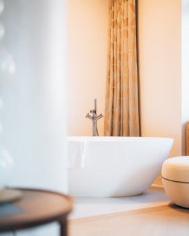 A modern, minimalist bathroom with a sleek white bathtub placed near a large window. Soft beige curtains hang next to the tub, complementing the gentle, neutral tones of the room. The edge of a circular light brown table is visible in the foreground, adding an elegant touch to the soft lighting in the space.