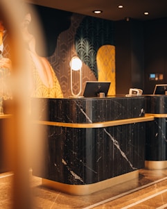 A modern and elegant hotel reception desk featuring a black marble finish with gold accents, complemented by a stylish lamp and computer on the countertop. The background displays a vibrant mural with geometric patterns and abstract art.