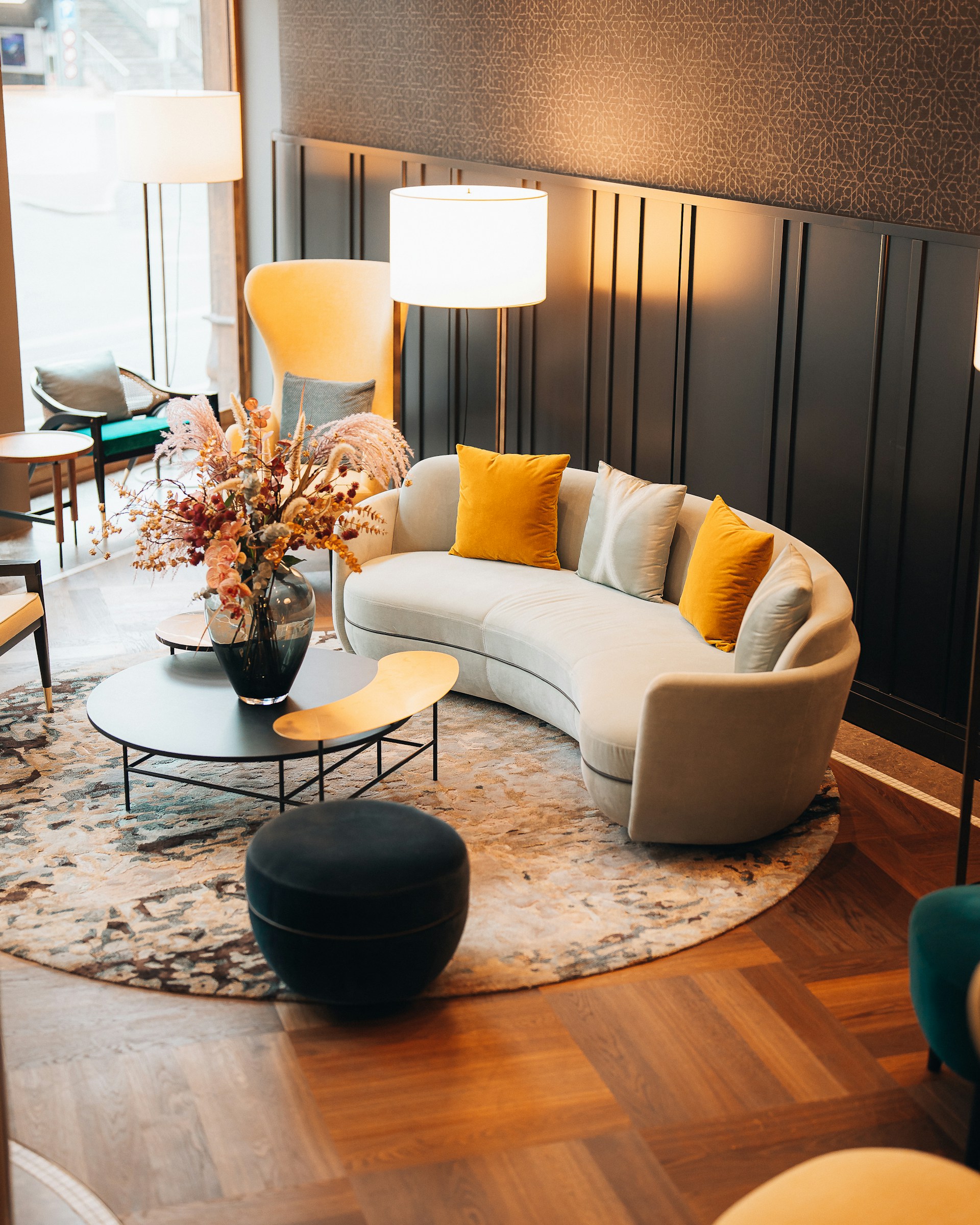 A modern living room featuring a curved, cream-colored sofa adorned with yellow and beige cushions. A large, decorative vase with dried flowers is placed on a round coffee table with a unique, asymmetric design. Two floor lamps provide warm lighting, and a large, patterned rug covers part of the wooden floor. The wall behind is lined with dark panels, adding to the cozy yet sophisticated ambiance.