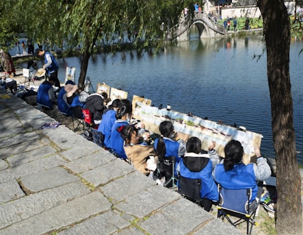 A group of people, primarily children, are sitting on portable chairs along a riverside. They are engaged in painting on canvas or paper set up in front of them. The scene includes a stone path leading down to the water, and the river reflects a stone arch bridge with people strolling across it. Lush greenery and trees surround the area, enhancing the peaceful ambiance.