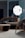 A modern interior setting featuring a stylish seating arrangement with a contemporary sofa, armchair, and a small table, all with sleek metal and dark fabric finishes. A large, illuminated photography umbrella is positioned behind the furniture, providing light and casting reflections on the shiny surfaces.