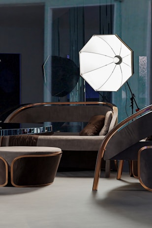 A modern interior setting featuring a stylish seating arrangement with a contemporary sofa, armchair, and a small table, all with sleek metal and dark fabric finishes. A large, illuminated photography umbrella is positioned behind the furniture, providing light and casting reflections on the shiny surfaces.