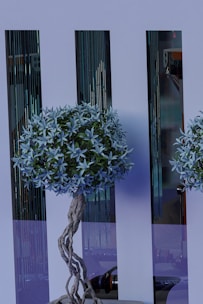 A decorative artificial tree with intricate twisted branches and lush foliage stands in front of sleek, reflective panels. The tree features a dense cluster of pale blue leaves and is complemented by the modern, stylish background.