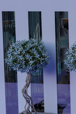 A decorative artificial tree with intricate twisted branches and lush foliage stands in front of sleek, reflective panels. The tree features a dense cluster of pale blue leaves and is complemented by the modern, stylish background.