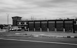 A large industrial building with multiple garage doors sits alongside a paved road. Several vehicles, including emergency response trucks, are parked in front of the building. The architecture features a control tower structure and the area appears deserted, with overcast skies adding to the somber atmosphere.