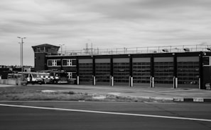 A large industrial building with multiple garage doors sits alongside a paved road. Several vehicles, including emergency response trucks, are parked in front of the building. The architecture features a control tower structure and the area appears deserted, with overcast skies adding to the somber atmosphere.