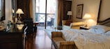 A cozy hotel room with a large unmade bed and stylish wooden furniture. There's a small sofa and a coffee table near a lamp. The room includes a work desk with a chair and a bottle of water on it. Two lamps on either side add warm lighting. Sheer curtains cover a set of sliding glass doors leading to a balcony with outdoor seating. The hardwood floor and elegant decor give the space a luxurious feel.