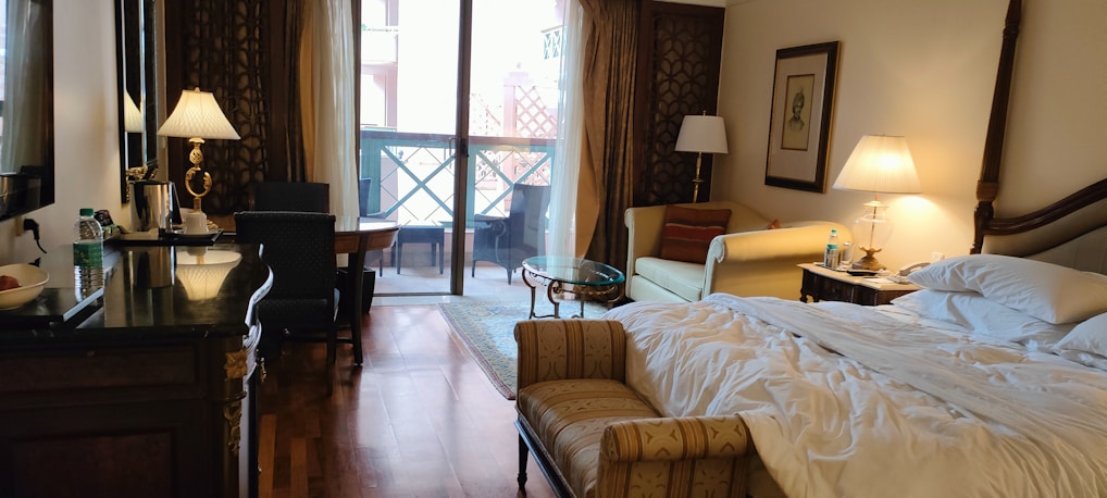 A cozy hotel room with a large unmade bed and stylish wooden furniture. There's a small sofa and a coffee table near a lamp. The room includes a work desk with a chair and a bottle of water on it. Two lamps on either side add warm lighting. Sheer curtains cover a set of sliding glass doors leading to a balcony with outdoor seating. The hardwood floor and elegant decor give the space a luxurious feel.