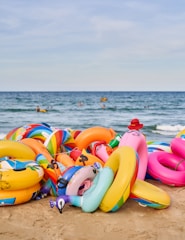 A pile of colorful inflatable rings and pool floats are lying on a sandy beach near the edge of the sea. In the background, the ocean waves are visible, and there are some people swimming and engaging in water activities.